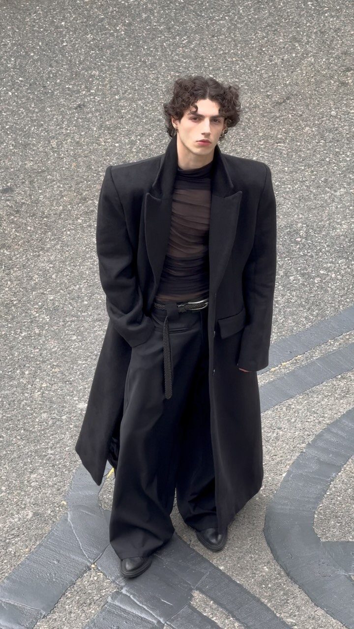 Man wearing a long black overcoat, sheer black top, and wide-leg black trousers.