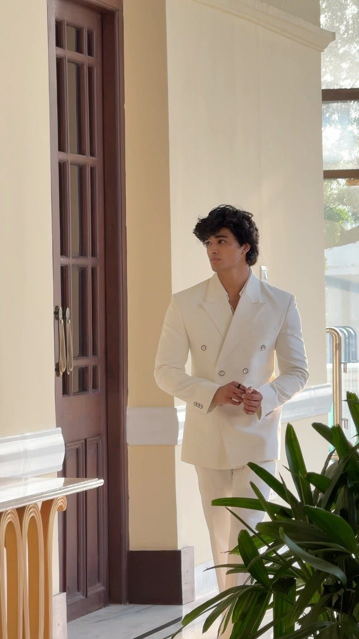 Man wearing a sophisticated double-breasted white suit standing in a sunlit hallway.