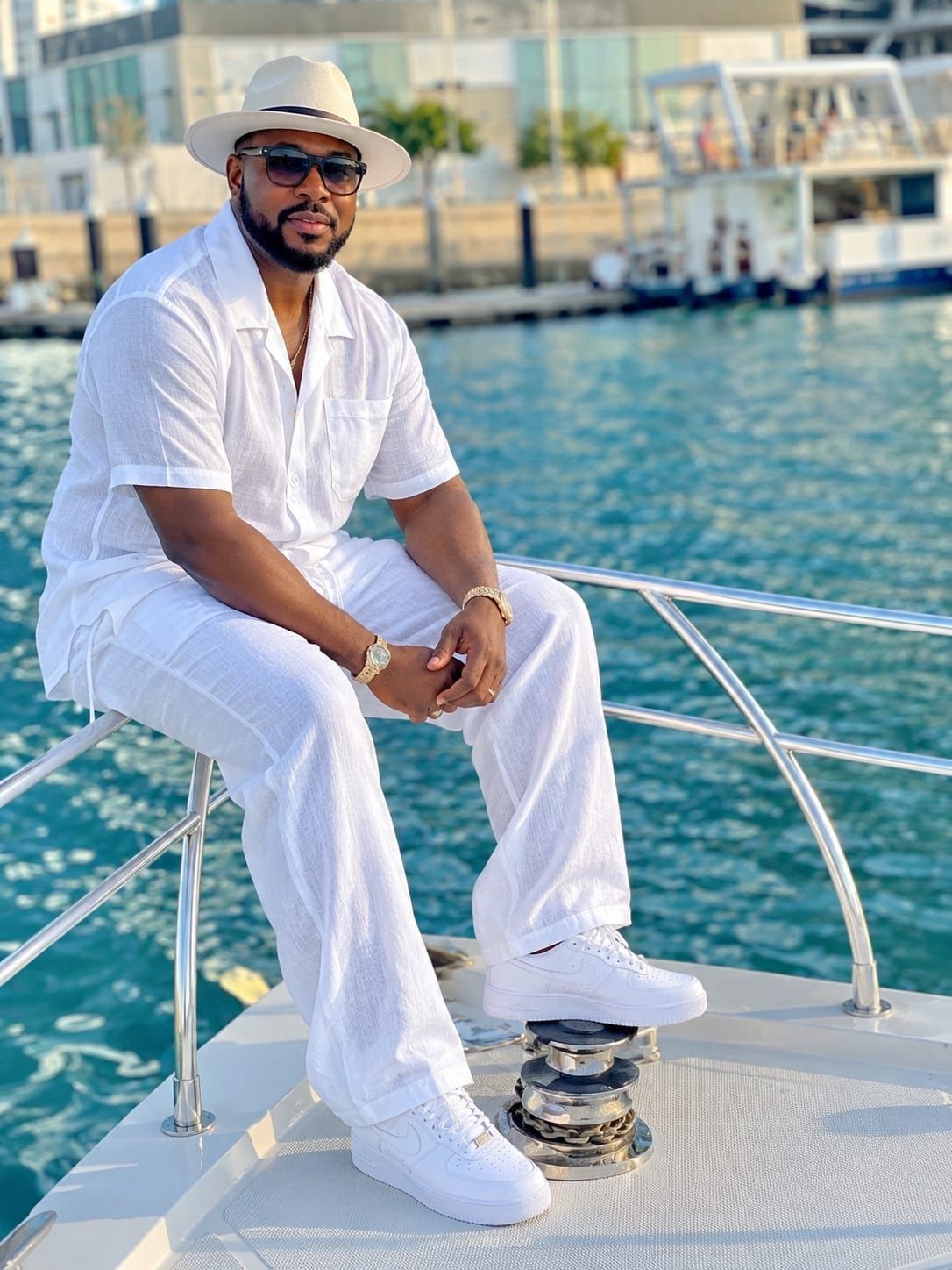 Man in all white outfit including hat and sneakers sitting on a boat.