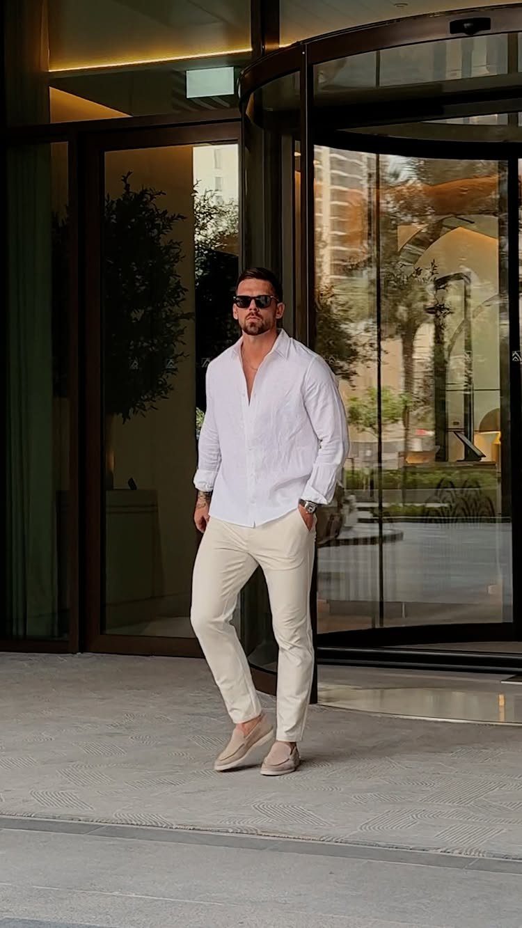 Man wearing a white long-sleeve shirt and white trousers standing by glass doors.