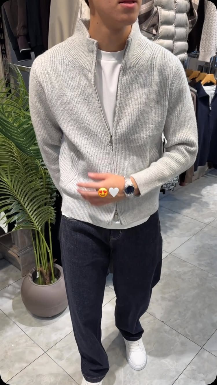Man wearing a light grey zip-up cardigan, dark trousers, and white sneakers.