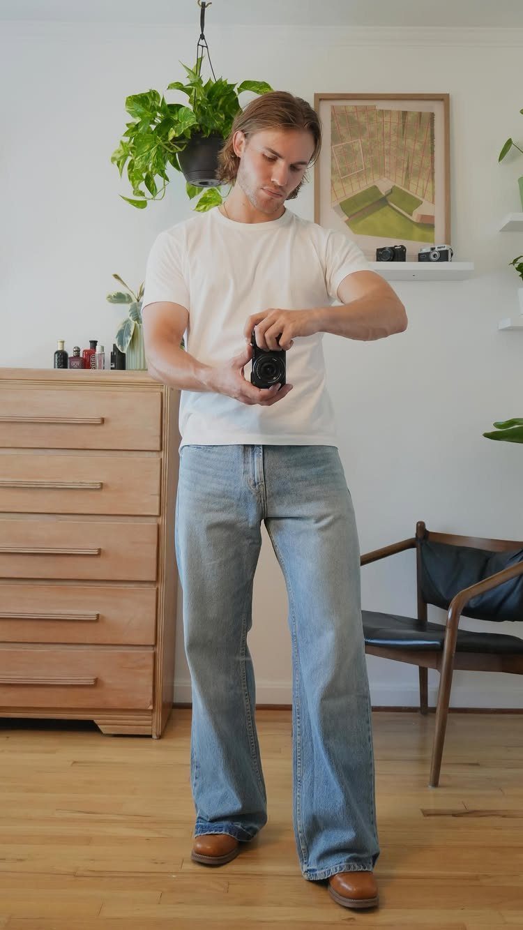 Man in white t-shirt and flared jeans holding a small black camera indoors.