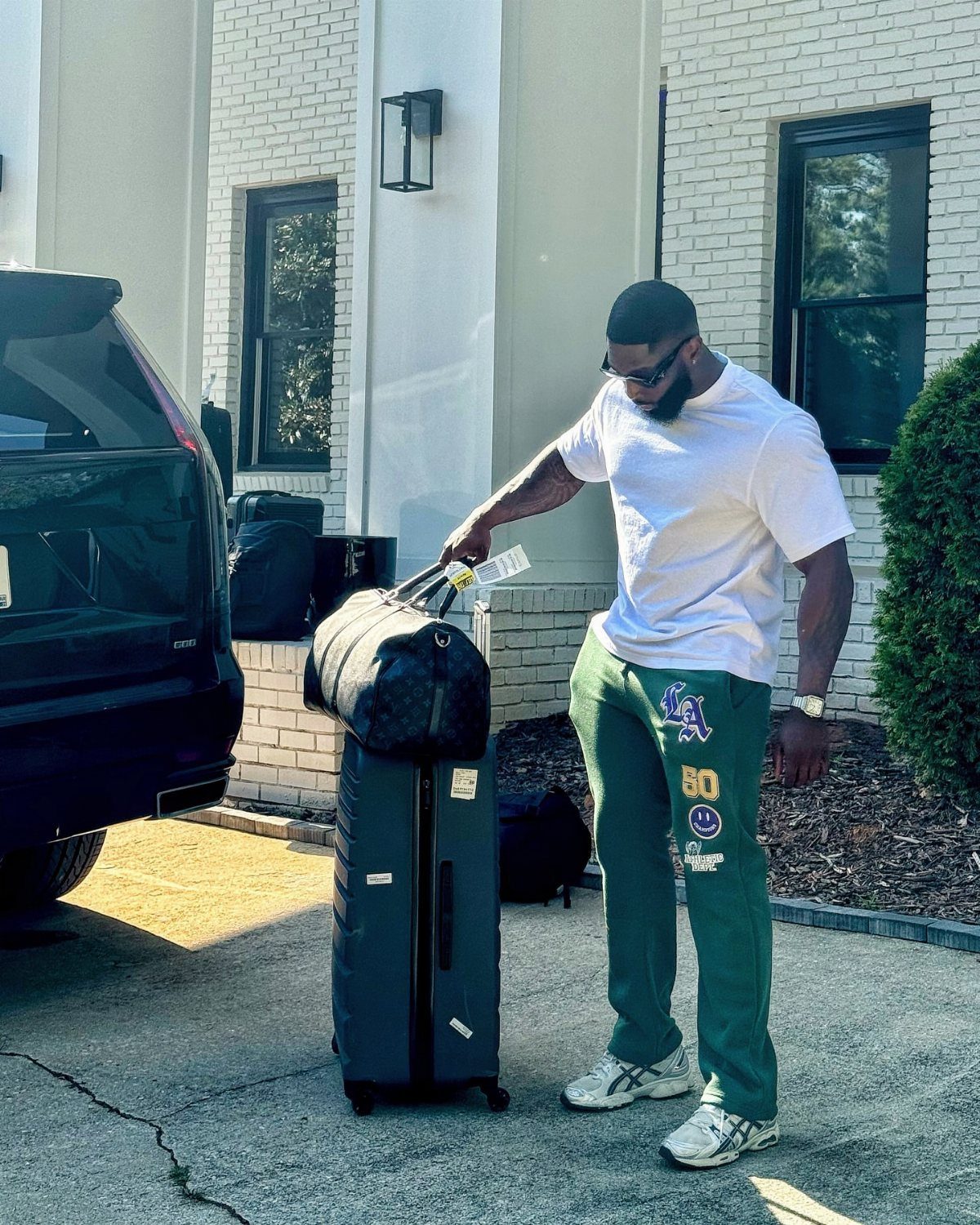 Man in a white t-shirt and green pants standing by a suitcase.