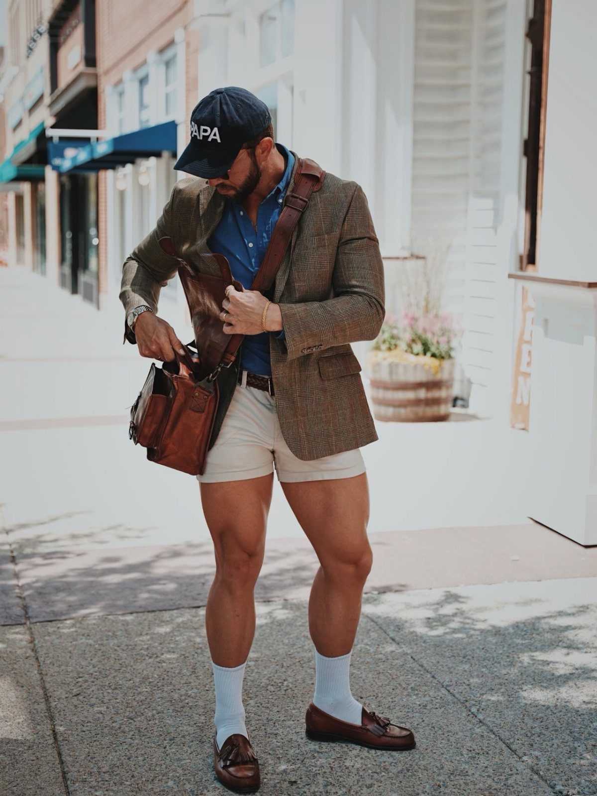 Man in brown patterned blazer and beige bermuda shorts holding a leather bag.