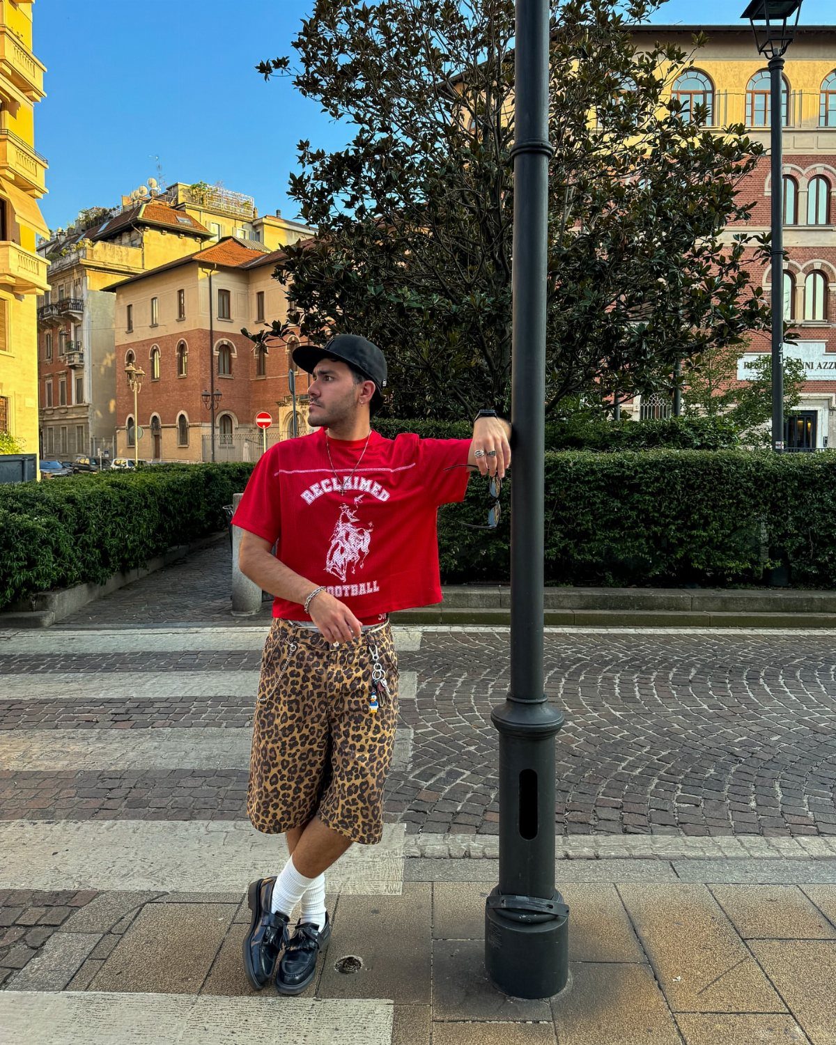 Man wearing red t-shirt and leopard print bermuda shorts leaning on lamp post