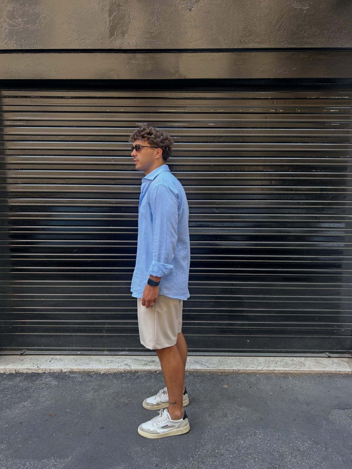 Man wearing a light blue linen shirt and beige bermuda shorts with white sneakers.