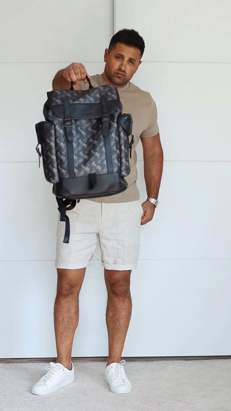 Man in tan t-shirt and off-white bermuda shorts holding a patterned backpack.