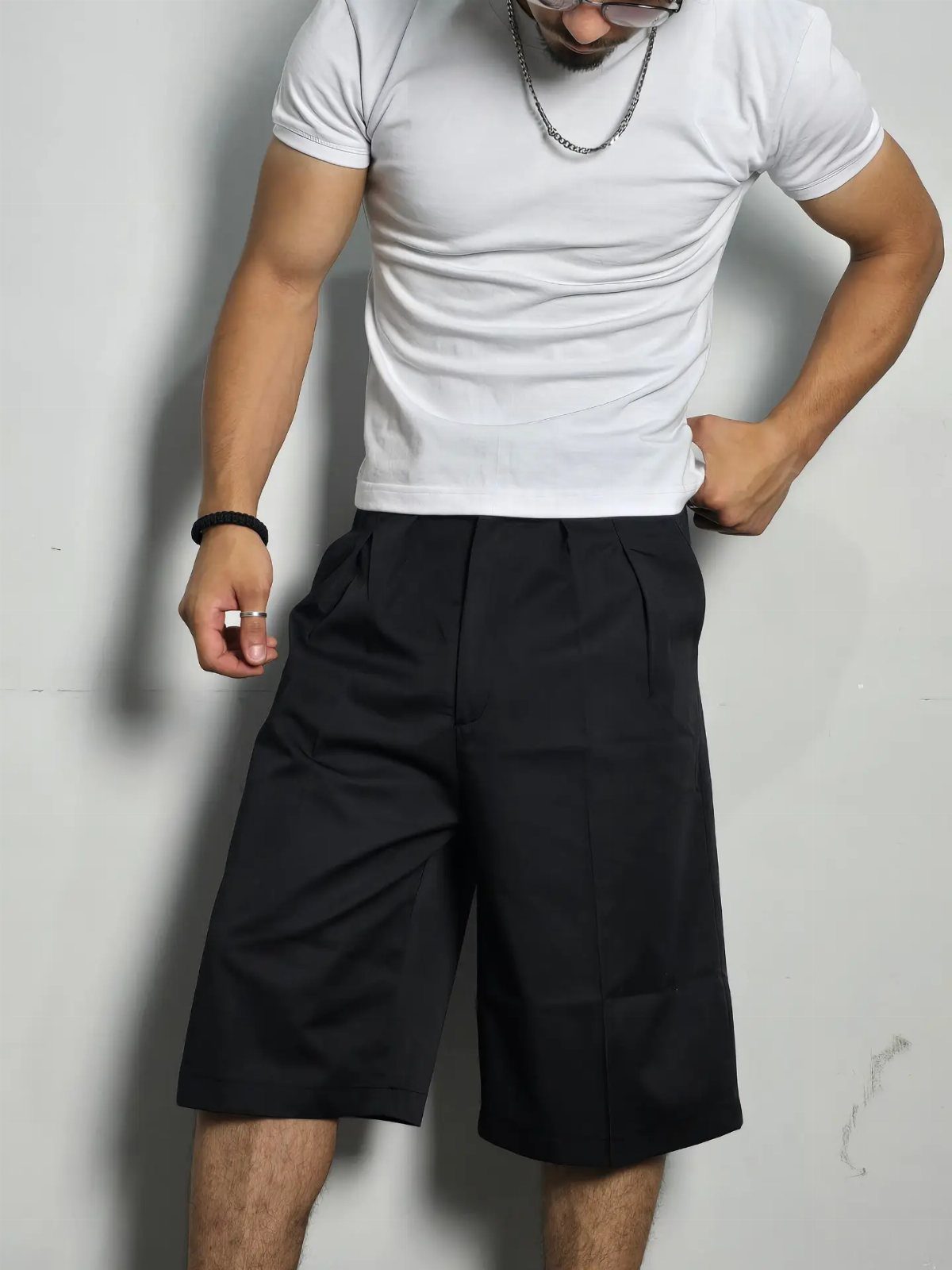 Man wearing white t-shirt and black wide-leg pleated bermuda shorts against grey wall