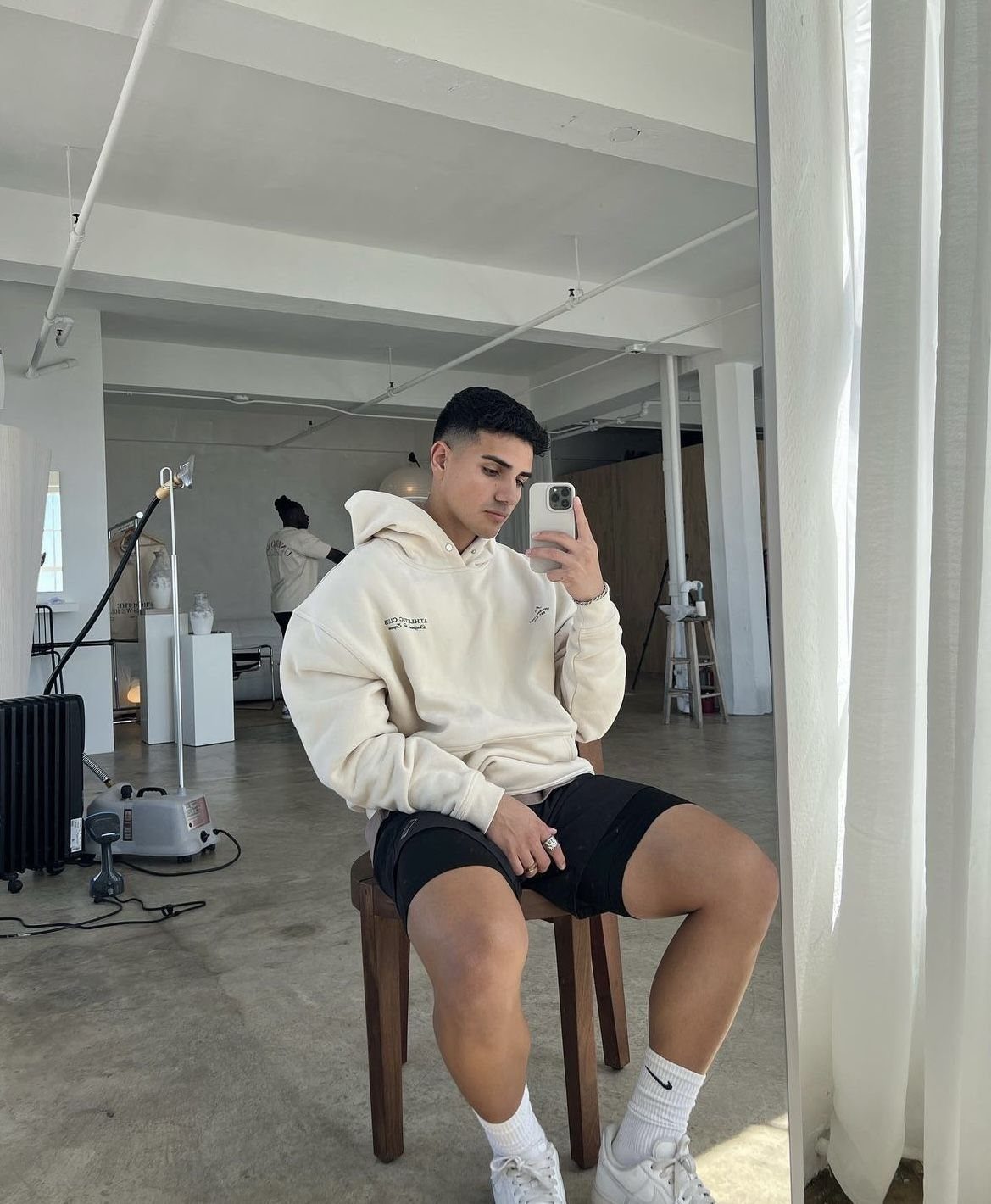 Man in cream hoodie and black biker shorts taking a mirror selfie in studio