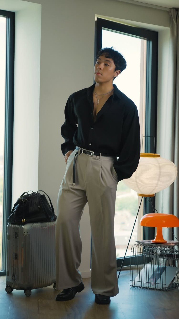Man in black button-down shirt and beige wide-leg trousers standing indoors with luggage.
