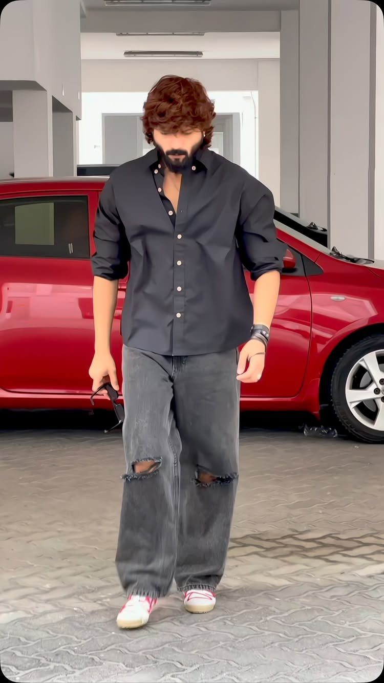 Man wearing a black button-down shirt and baggy grey ripped jeans by a red car