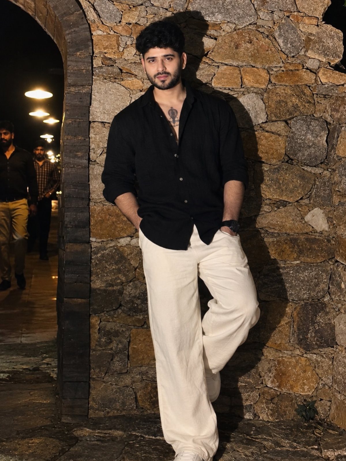 Man wearing a black button-down shirt and wide-leg white trousers against stone wall