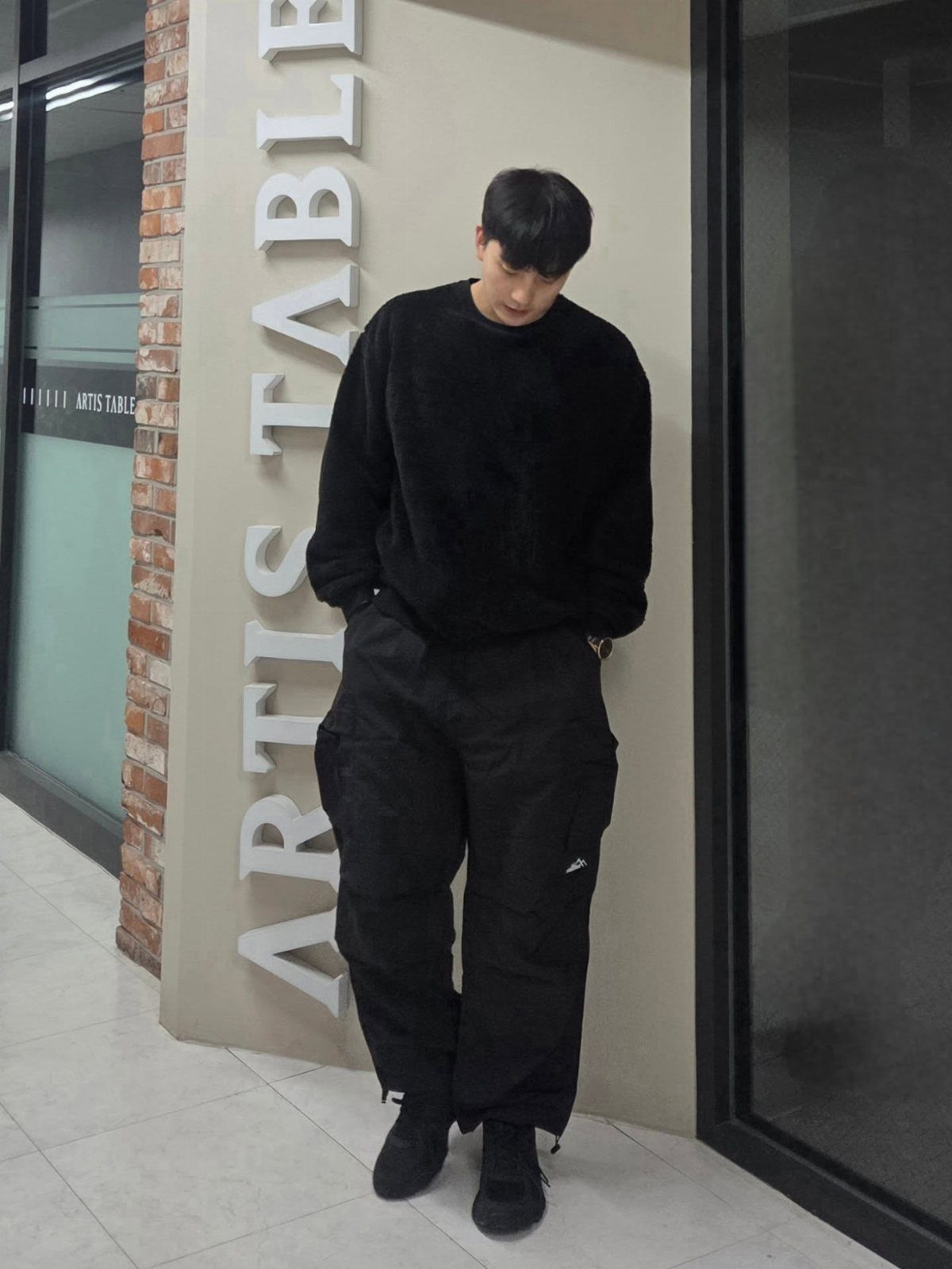 Man wearing a black fuzzy sweater and black cargo pants standing indoors.
