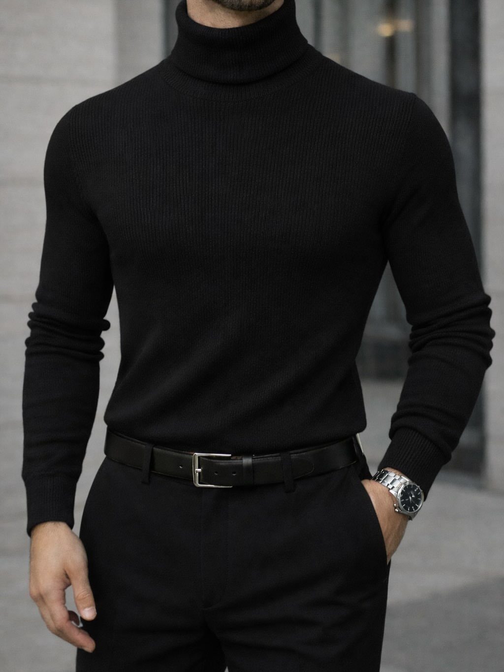 Man wearing a black ribbed turtleneck sweater tucked into black trousers with belt.