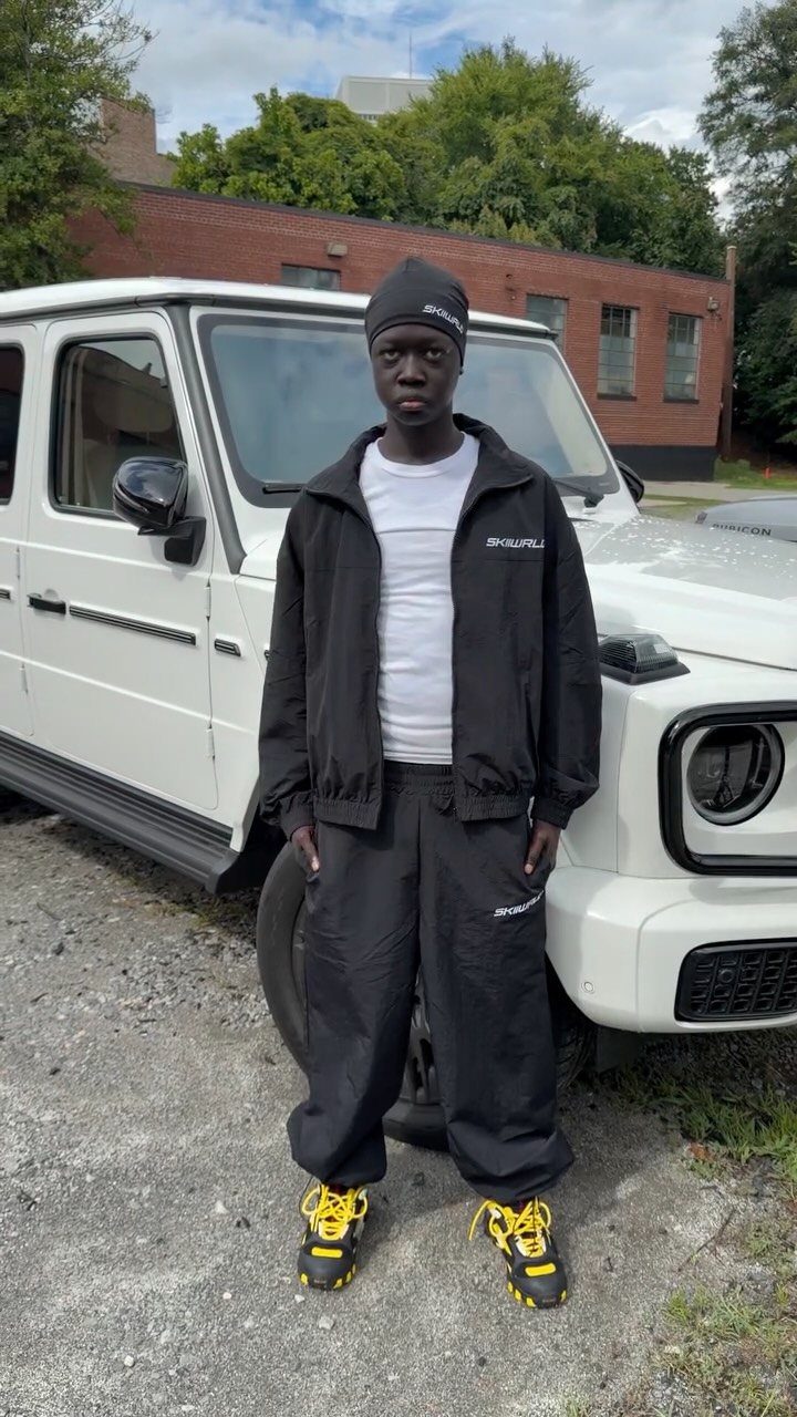 Person wearing baggy black trousers, black jacket, and yellow-laced sneakers by a white SUV.