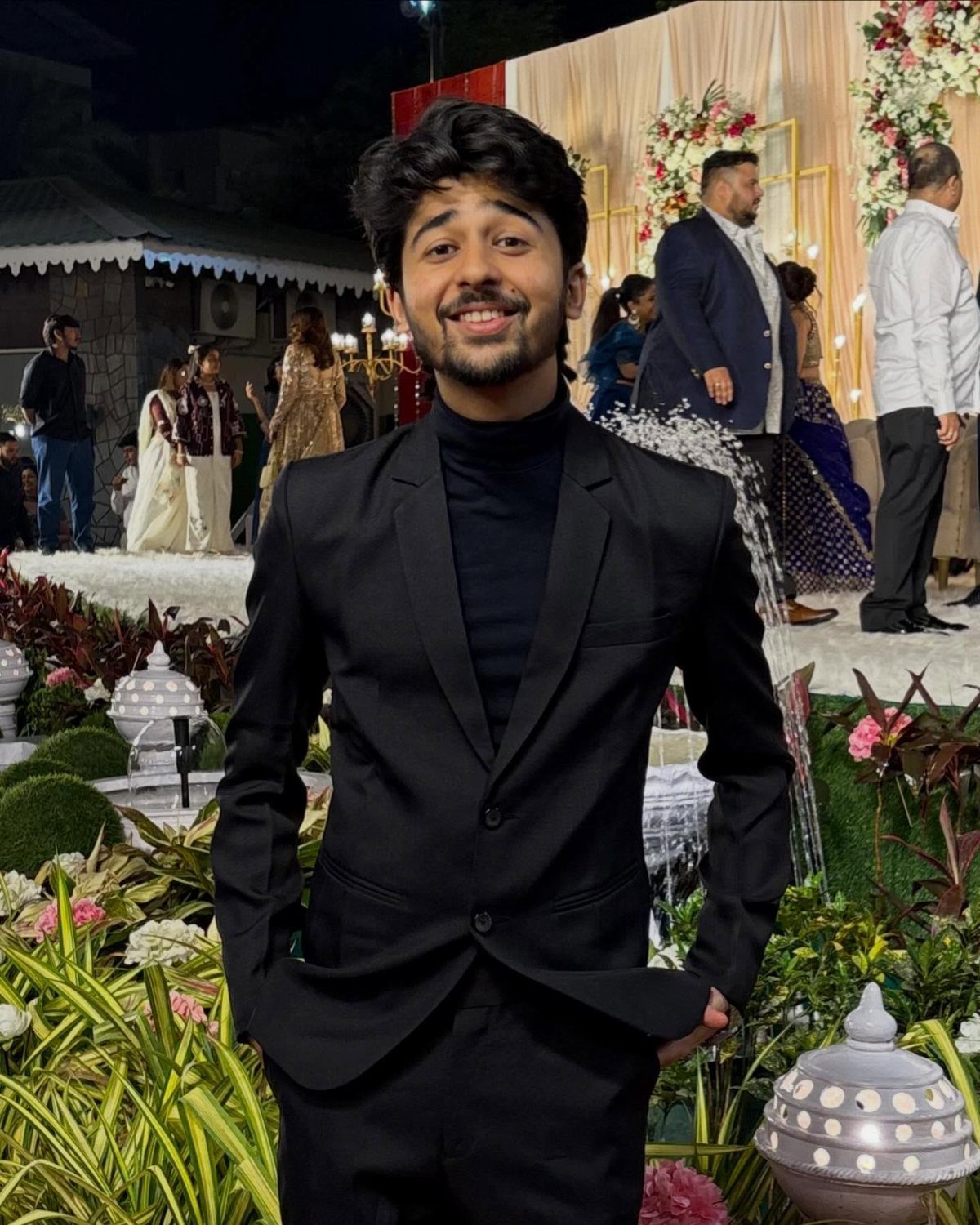 Man wearing a black turtleneck under a black tailored dinner jacket in a garden.