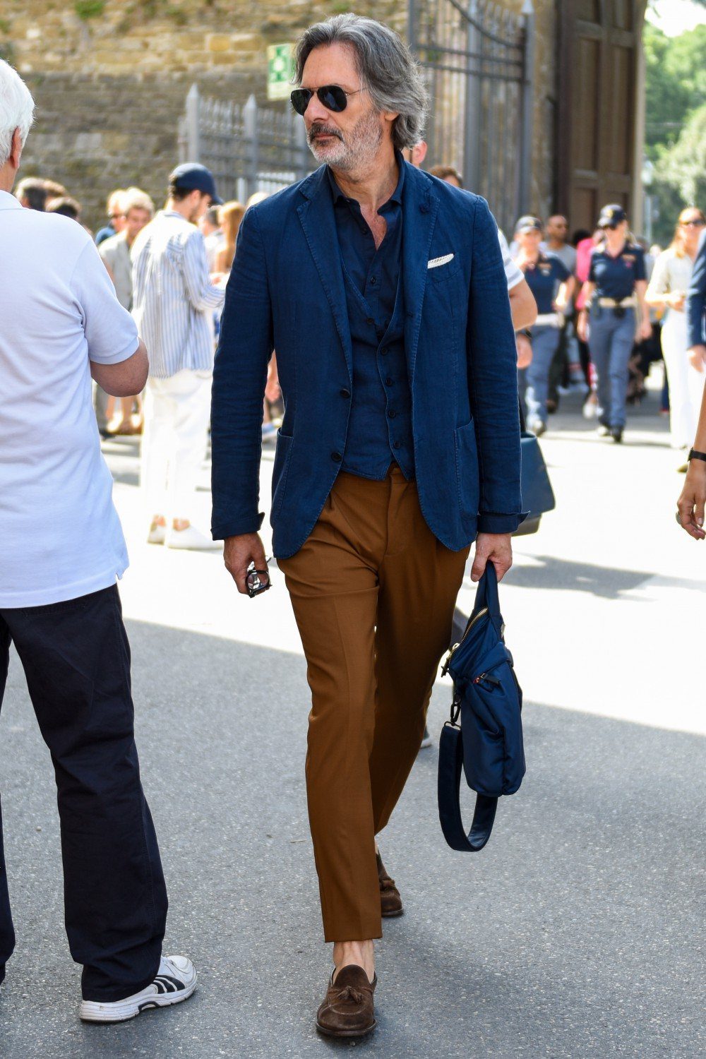 Man wearing navy blazer, blue shirt, and brown pants walking on a street
