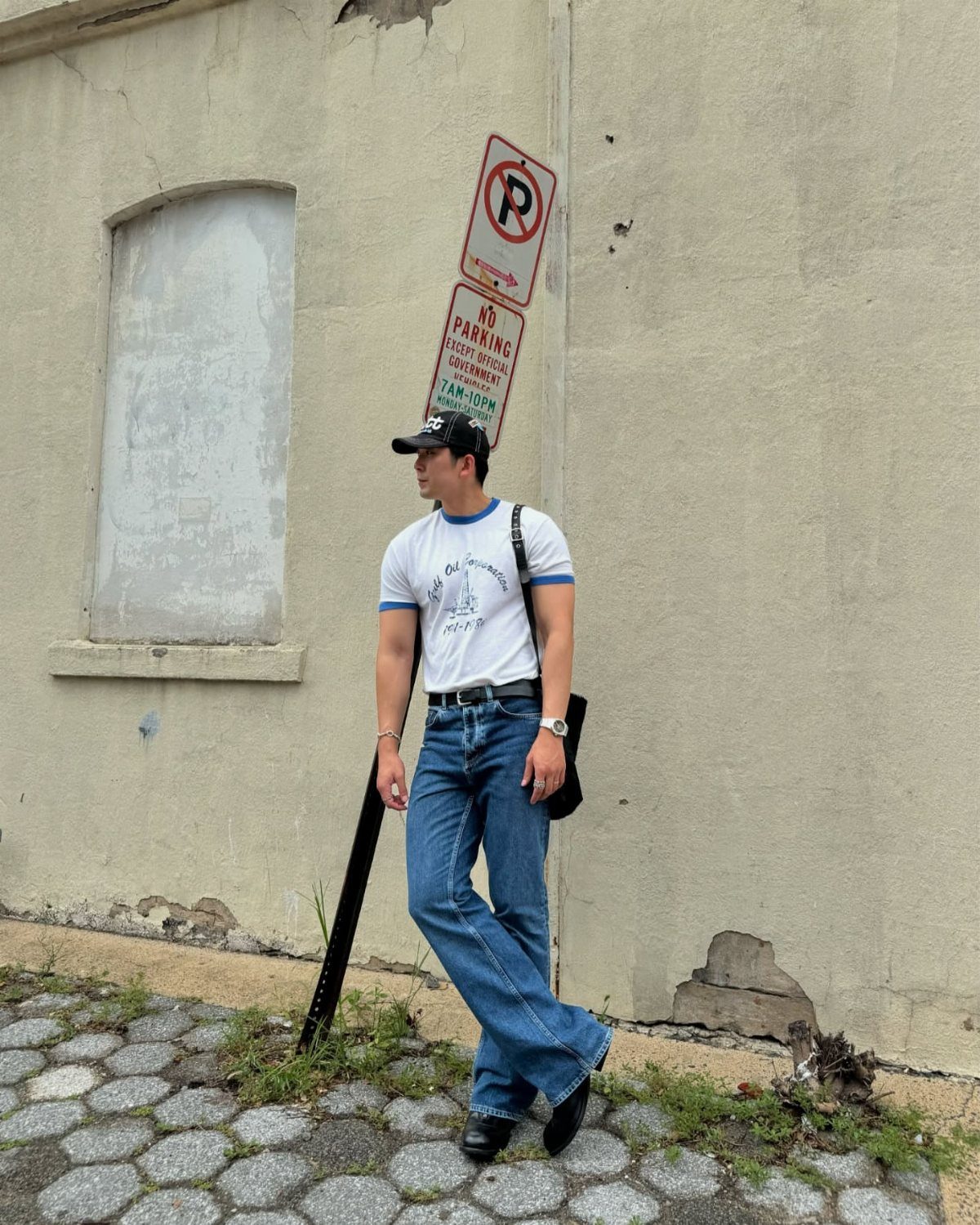 Man wearing a white graphic t-shirt and blue bootcut jeans standing by a wall.
