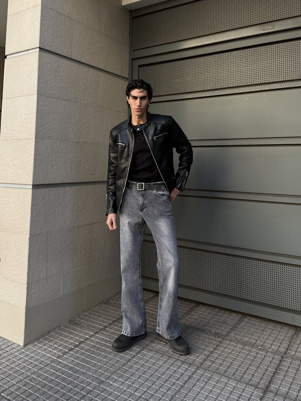 Man wearing a black leather jacket and grey bootcut jeans standing by a wall