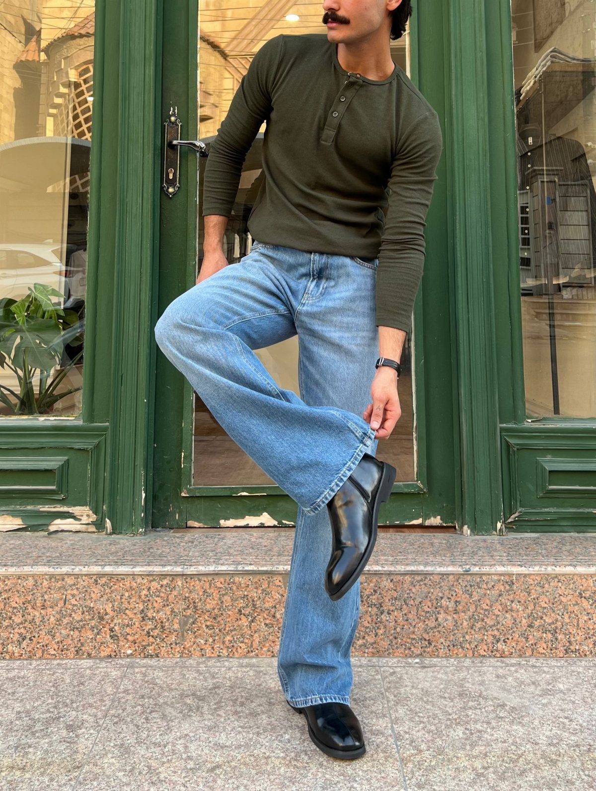 Man in olive henley and light blue bootcut jeans posing by green door