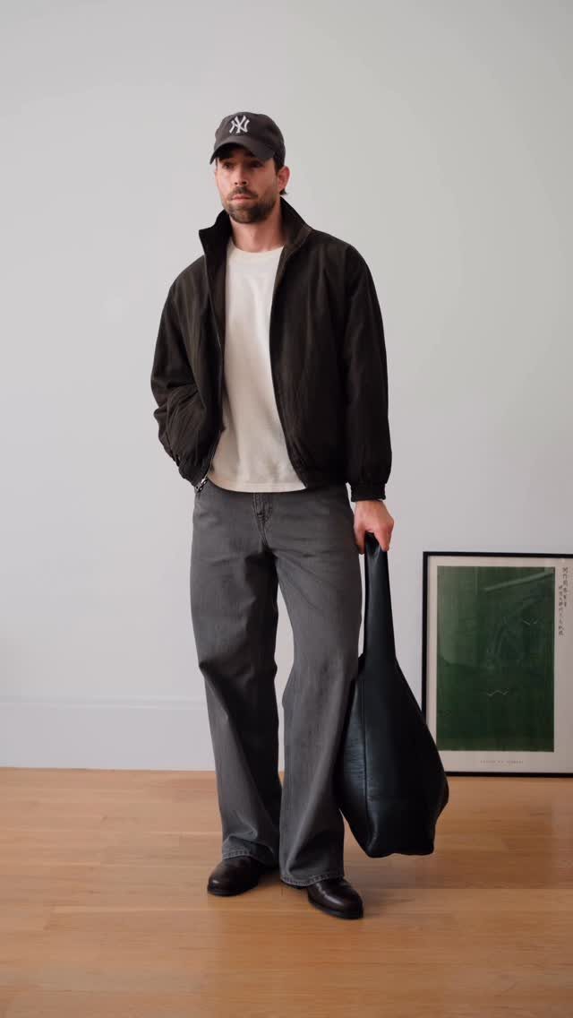 Man wearing a brown jacket and grey trousers standing in a minimalist room.