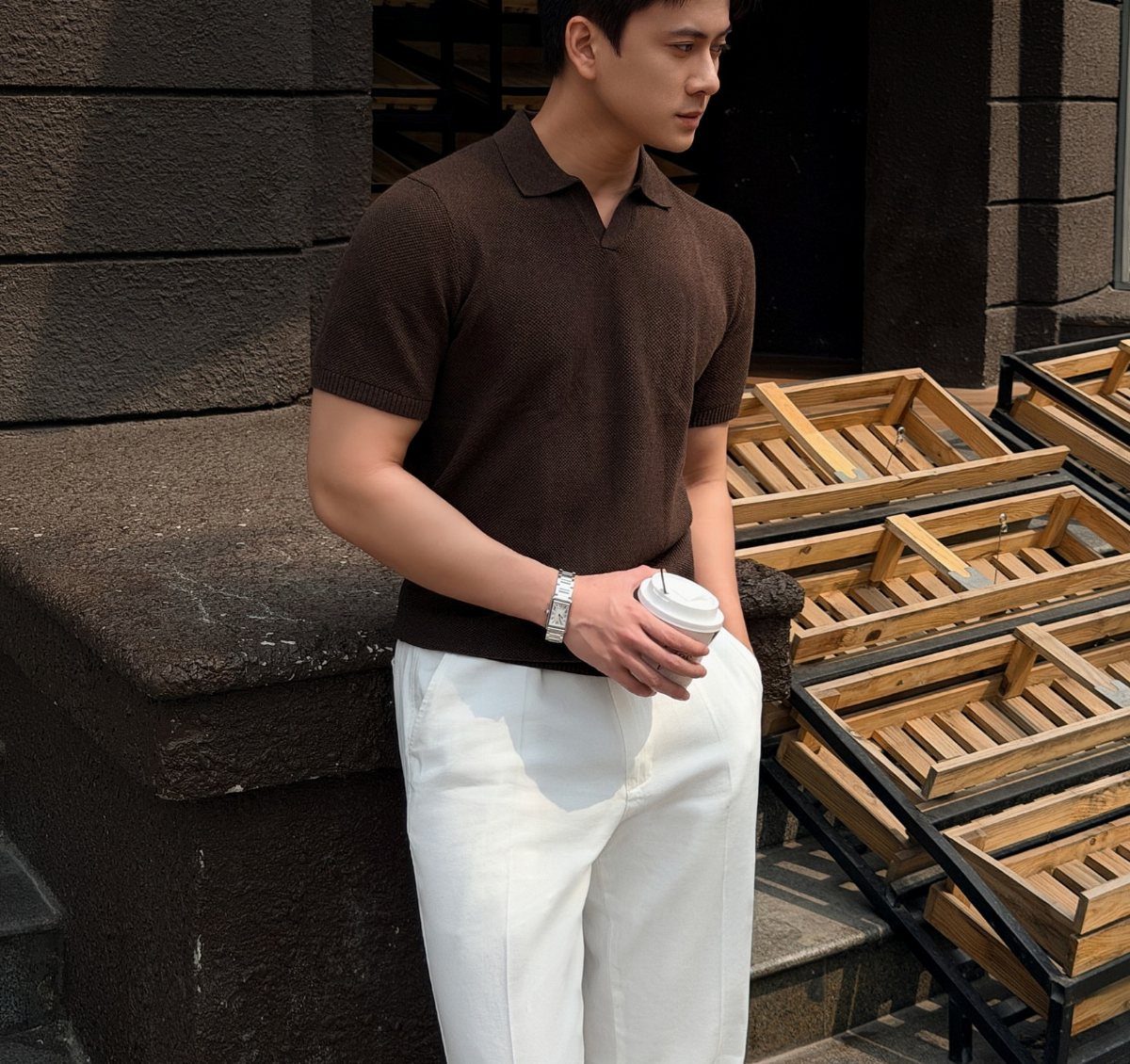 Man in a brown knit polo shirt and white trousers holding a coffee cup.