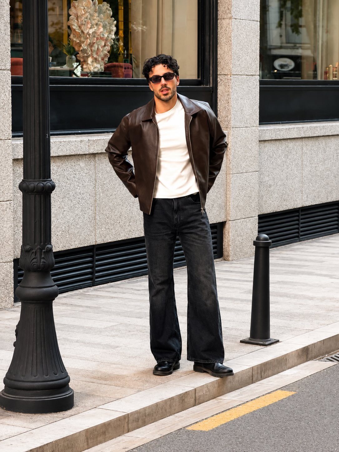 Man wearing a brown leather jacket, white t-shirt, and wide black jeans outdoors.