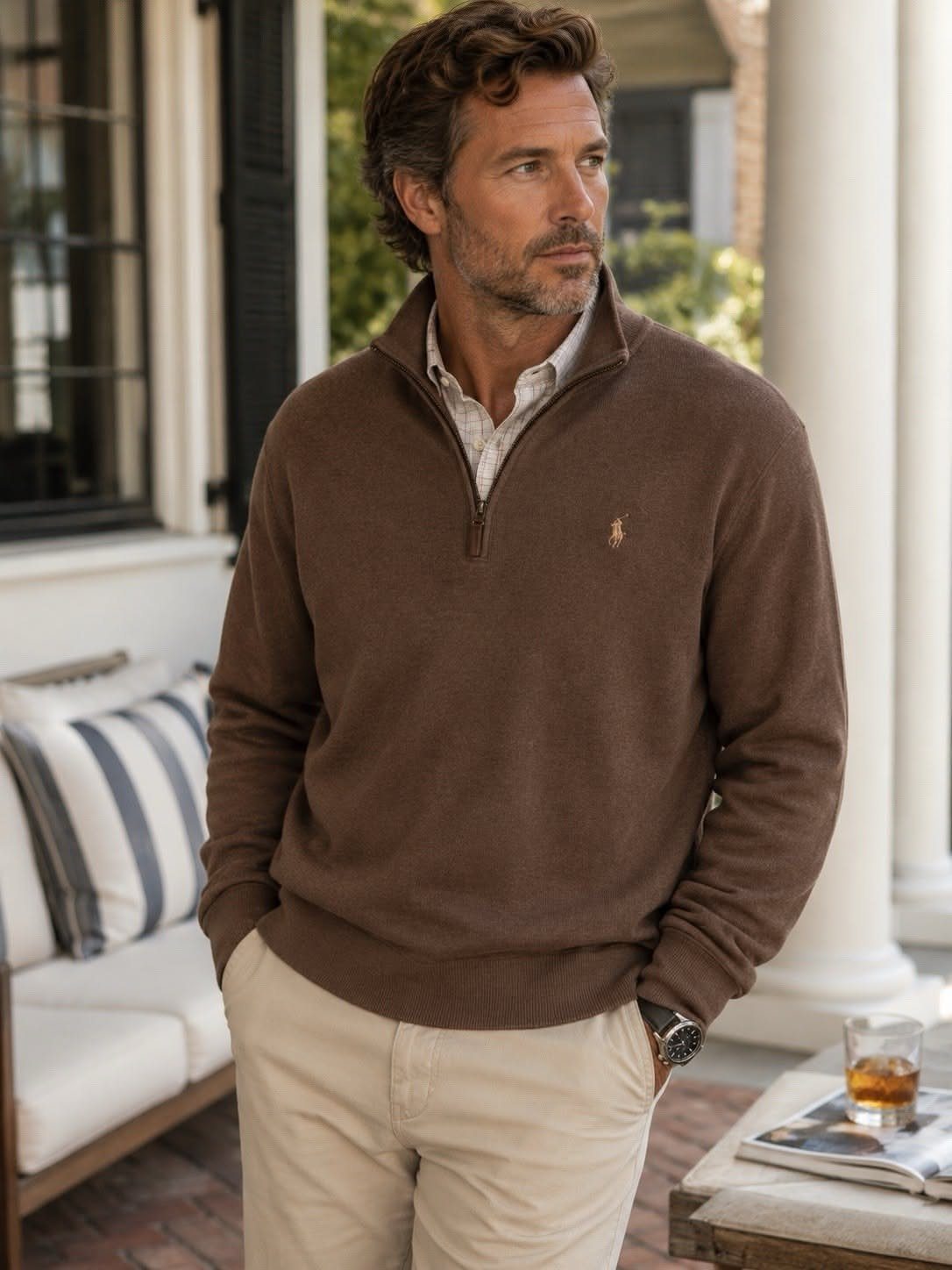 Man wearing a brown quarter-zip sweater and beige trousers standing on a porch.
