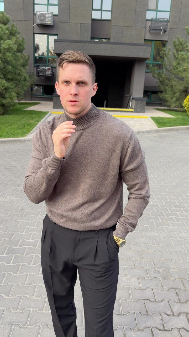 Man wearing a brown turtleneck sweater and black trousers standing outdoors near a building