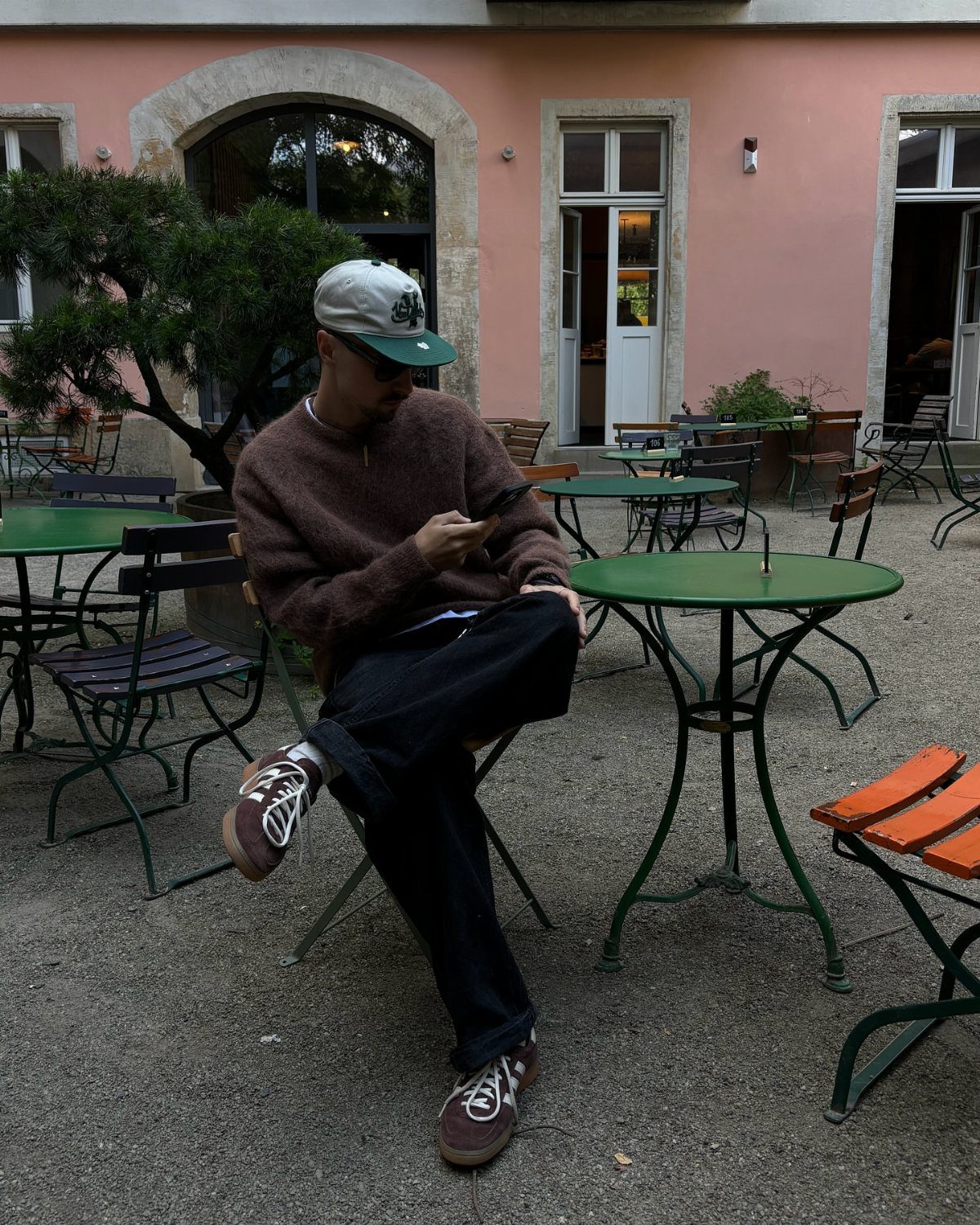 Man in brown sweater and dark denim pants sitting at outdoor cafe table