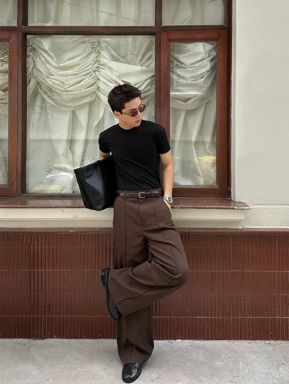 Man in black t-shirt and wide-leg brown trousers leaning against a tiled wall.