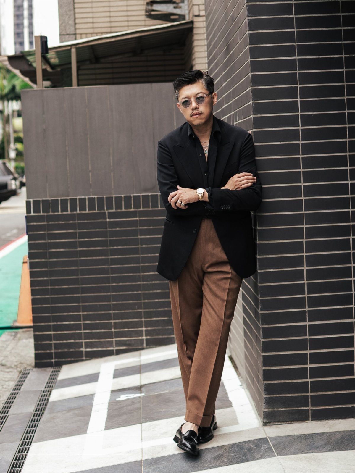 Man in black blazer and brown trousers leaning against a dark brick wall.