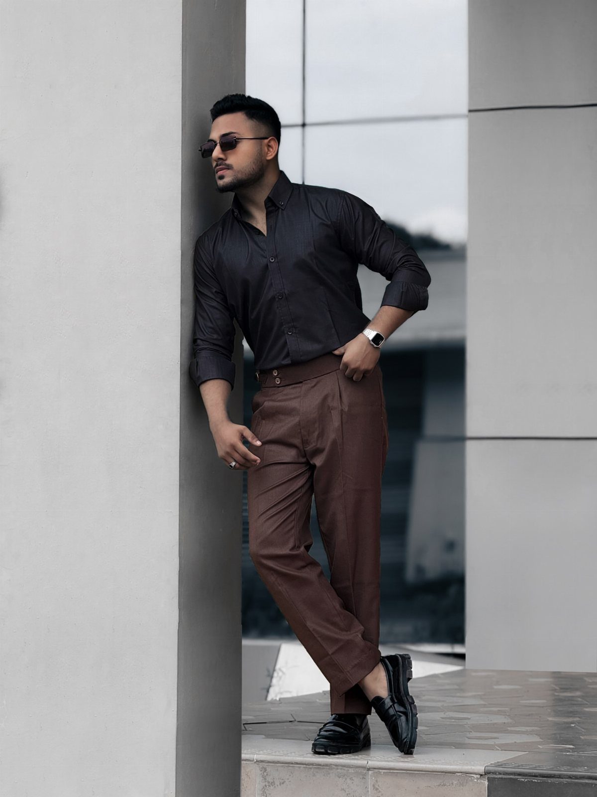 Man in black shirt and brown trousers leaning against a white wall outdoors.