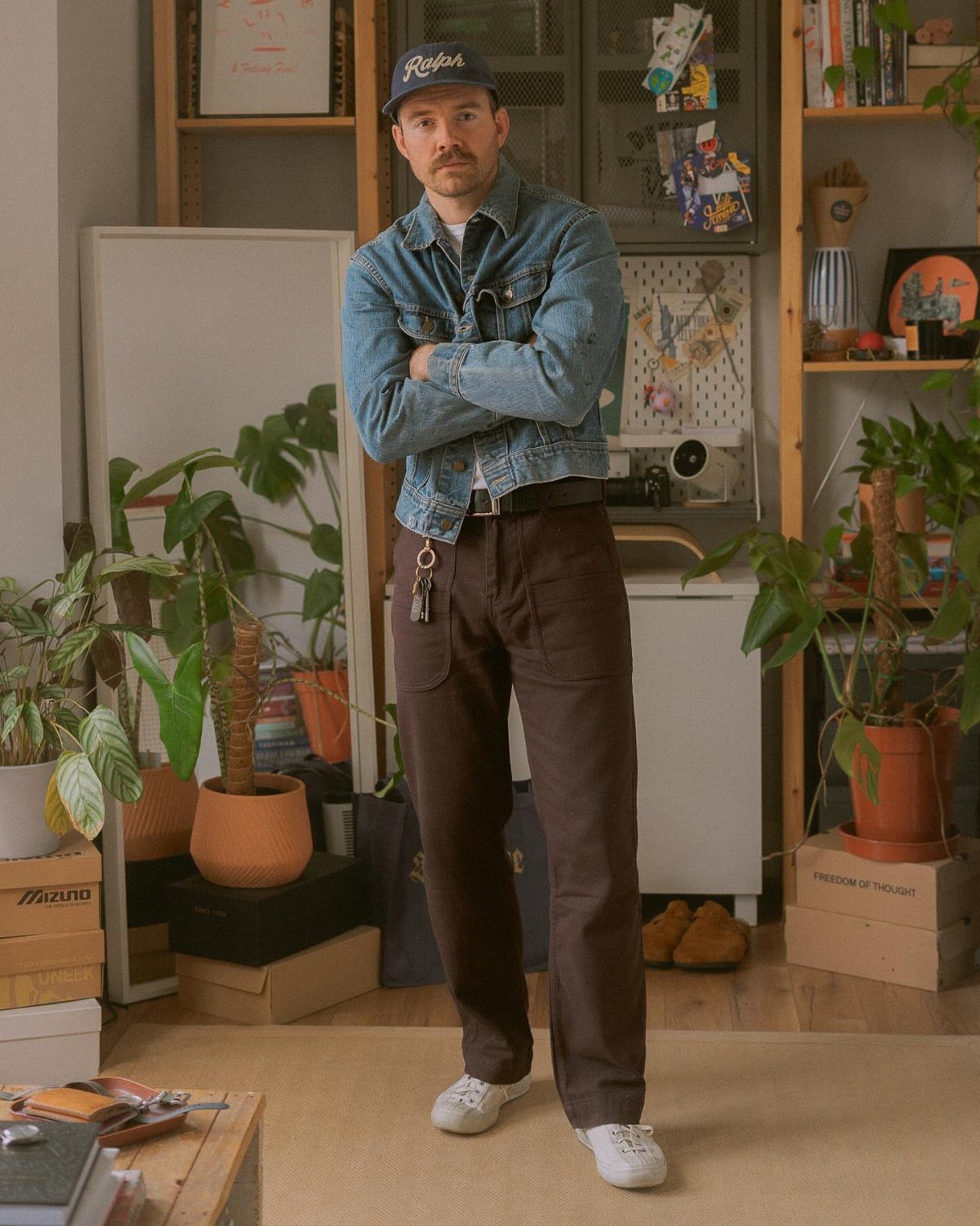 Man wearing a denim jacket, blue cap, brown trousers, and white sneakers indoors.