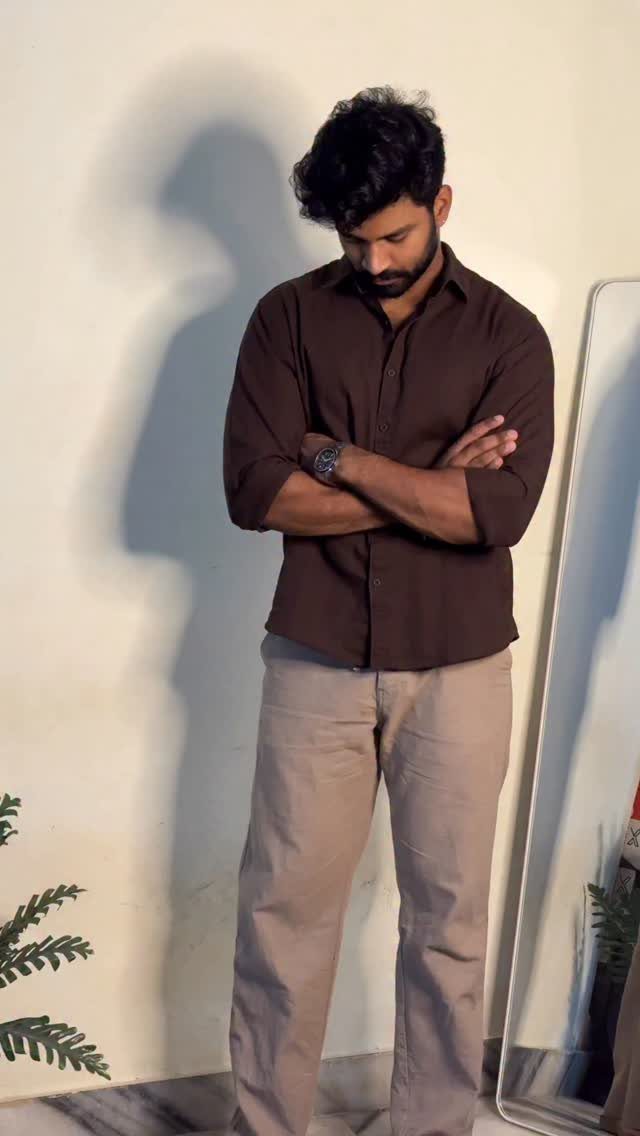 Man wearing a dark brown shirt and light brown trousers standing against a wall