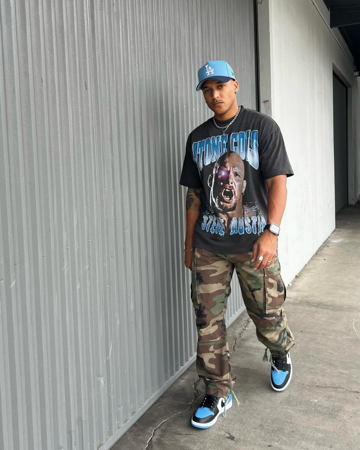 Man wearing blue cap, black graphic tee, camo cargo pants, and blue sneakers.