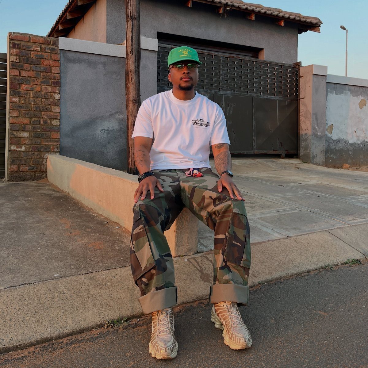 Man wearing green camo pants, white t-shirt, and a green baseball cap sitting outdoors.