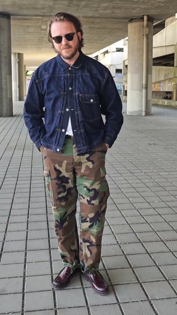 Man wearing a denim jacket, camouflage pants, and burgundy shoes on a tiled plaza.