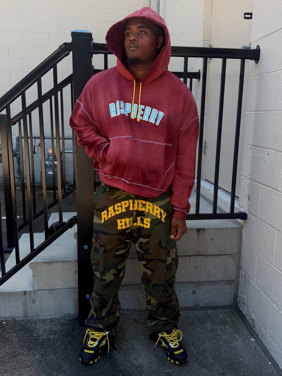 Man wearing a maroon hoodie and camouflage cargo pants standing by a railing.