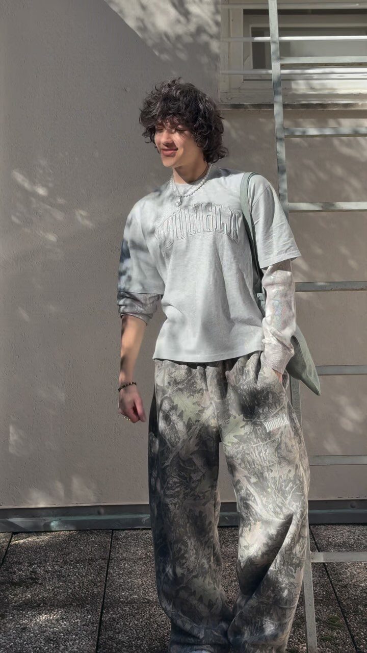 Young man wearing a grey t-shirt and wide-leg camouflage pants standing outdoors.