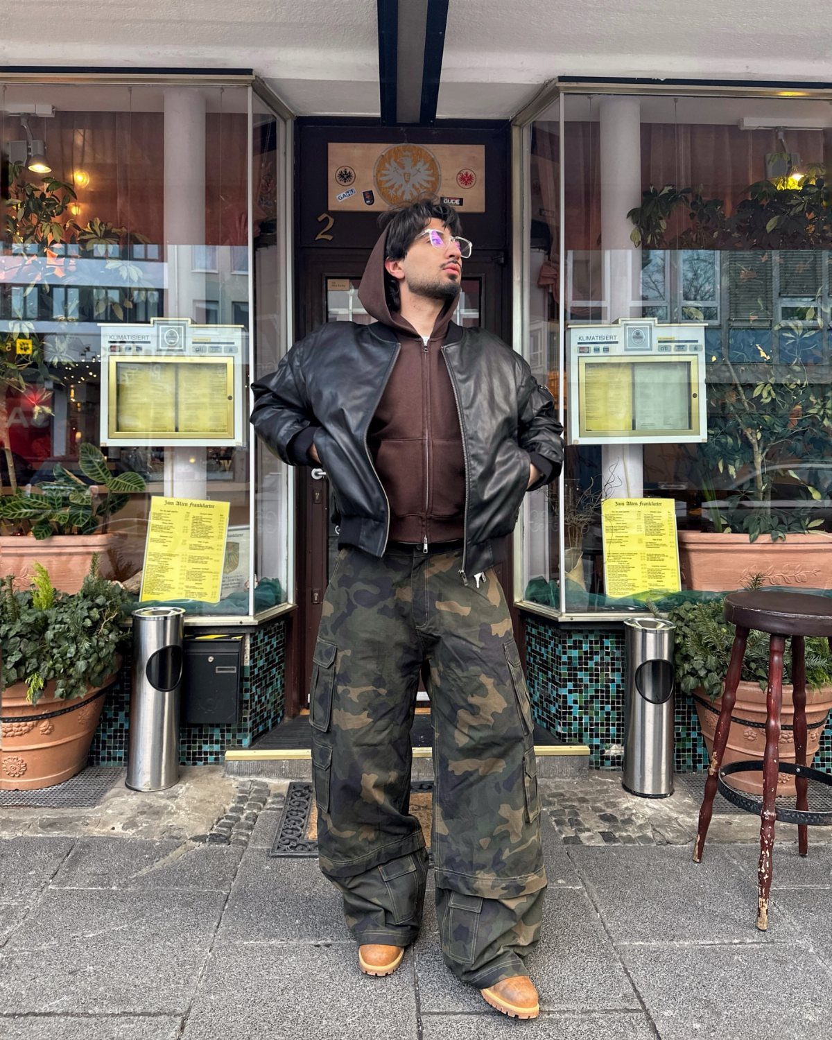 Man wearing a black leather jacket, brown hoodie, and baggy camo cargo pants.