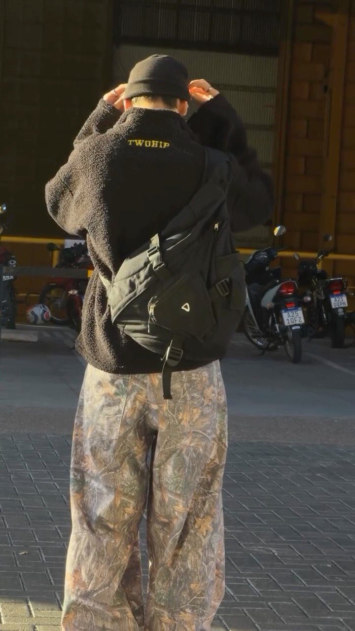 Person wearing camouflage pants, a black fleece jacket, and a black sling bag.