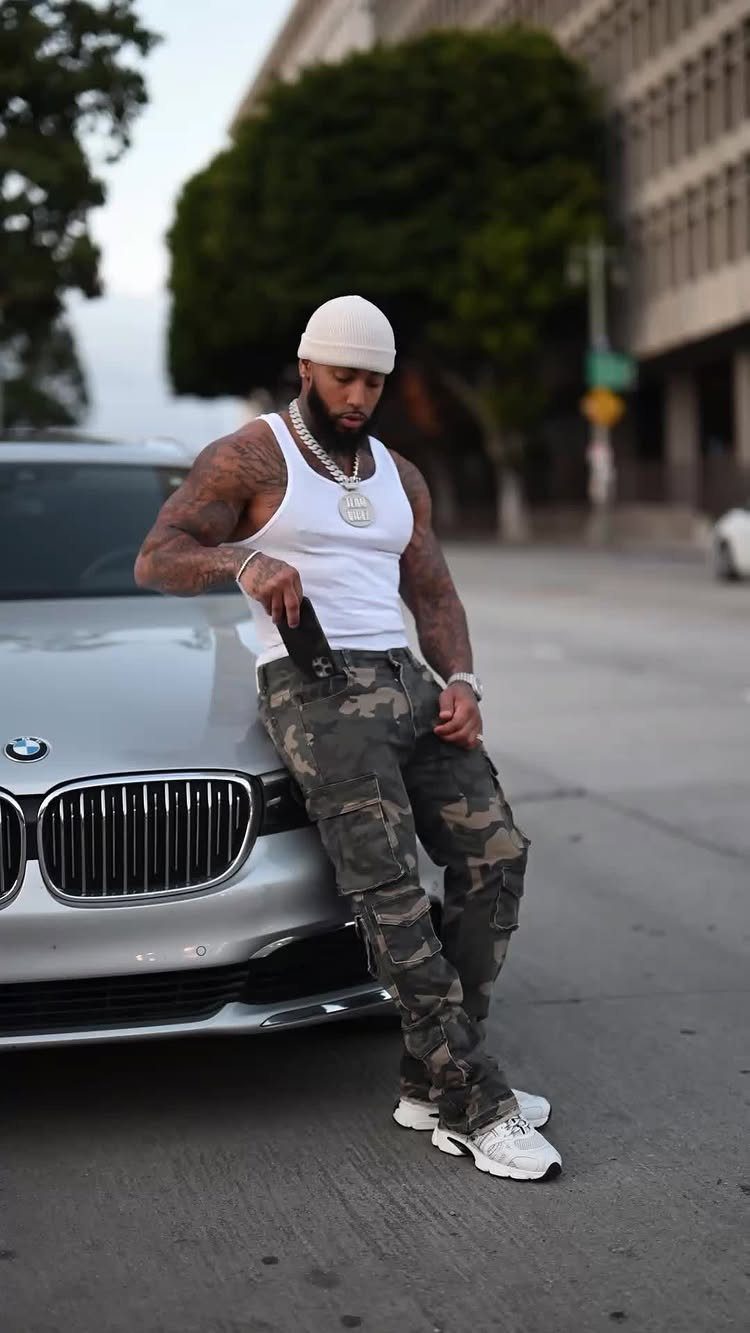 Man in camouflage cargo pants and white tank top leaning on silver car
