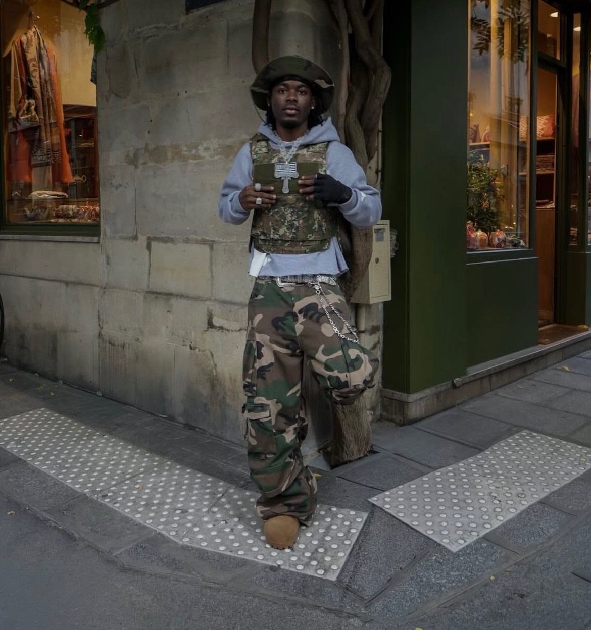 Man wearing a grey hoodie, tactical vest, and camouflage pants on a city street.
