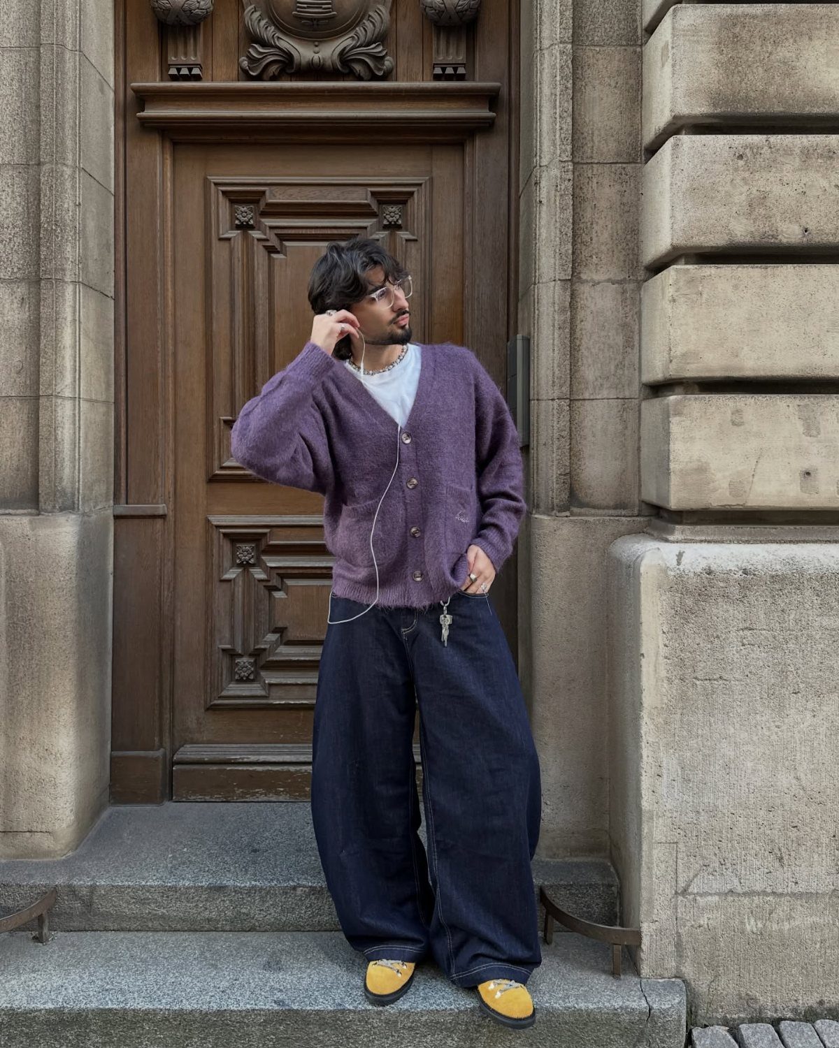 Man wearing a purple button-up cardigan and wide-leg dark blue jeans by a door.