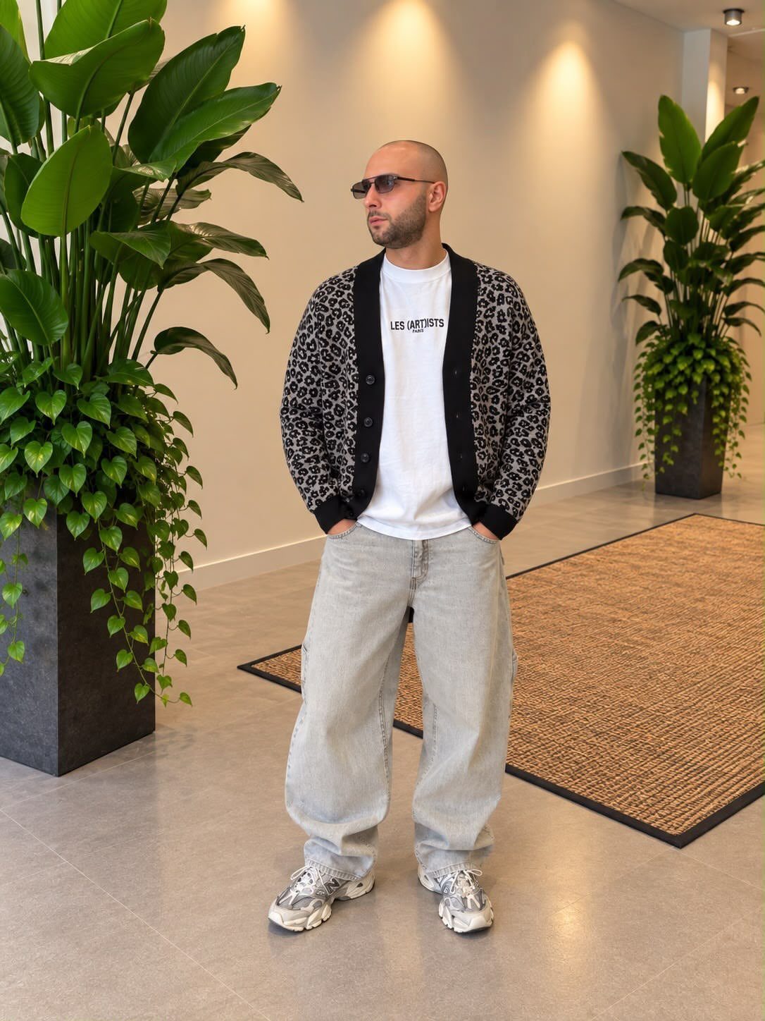 Man wearing a patterned cardigan, white t-shirt, and wide-leg grey jeans indoors.