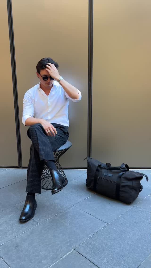 Man in white shirt and pinstriped trousers sitting with a black travel bag.