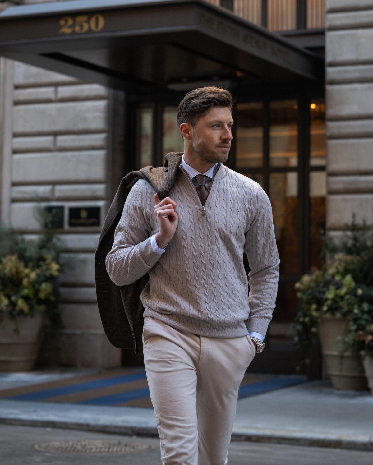 Man wearing a grey cable-knit sweater and cream trousers standing outside a building.