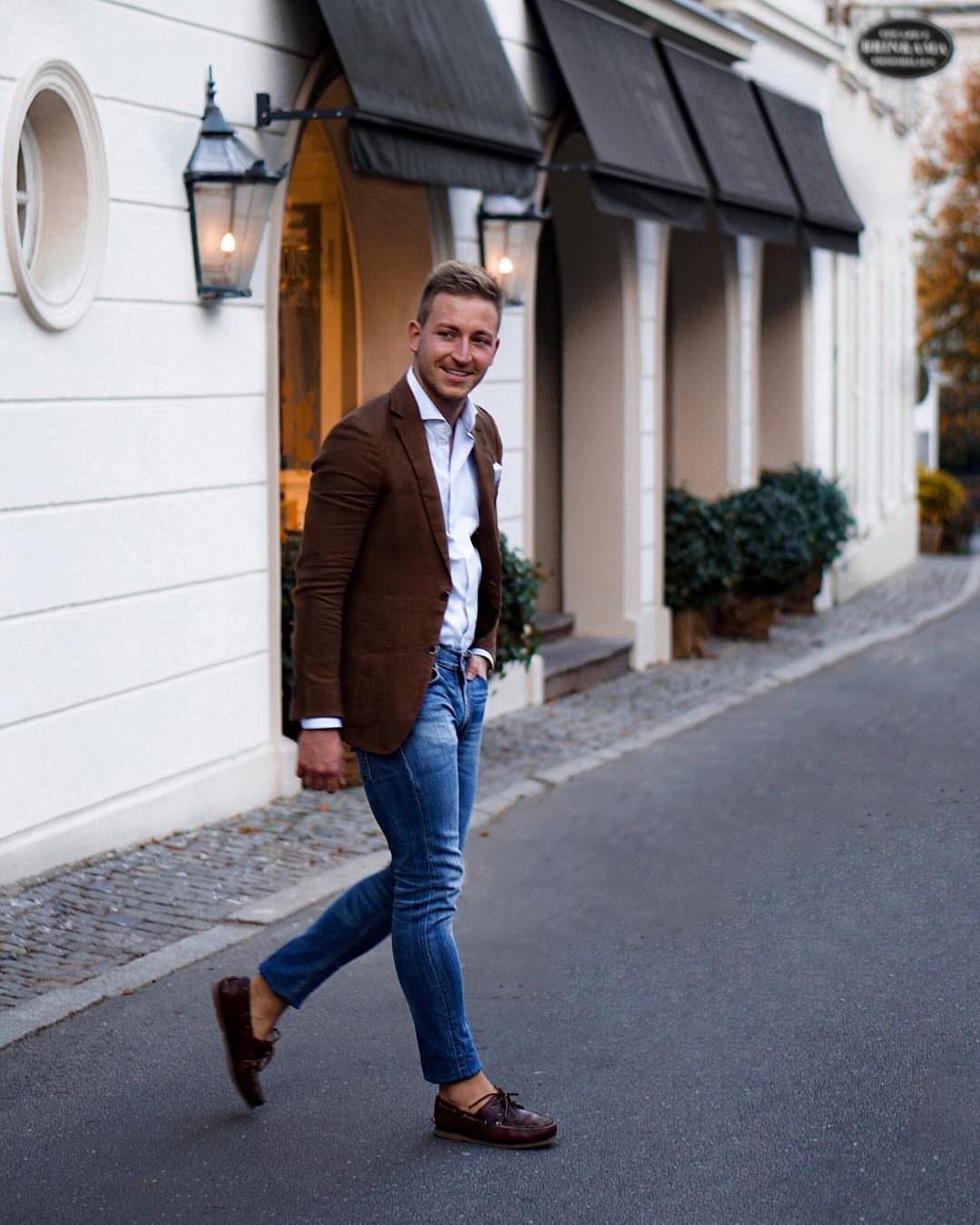 Man wearing a brown blazer and blue jeans walking on a city street.