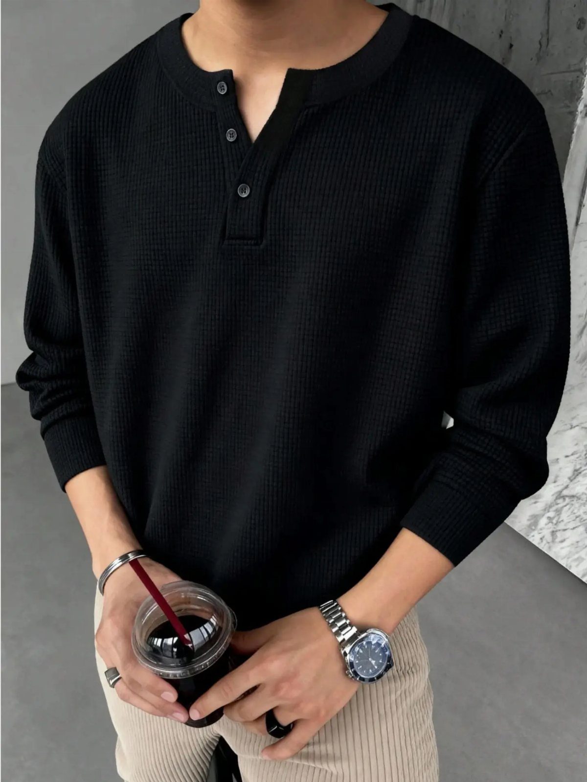 Man wearing a black waffle-knit henley shirt and beige trousers holding a drink